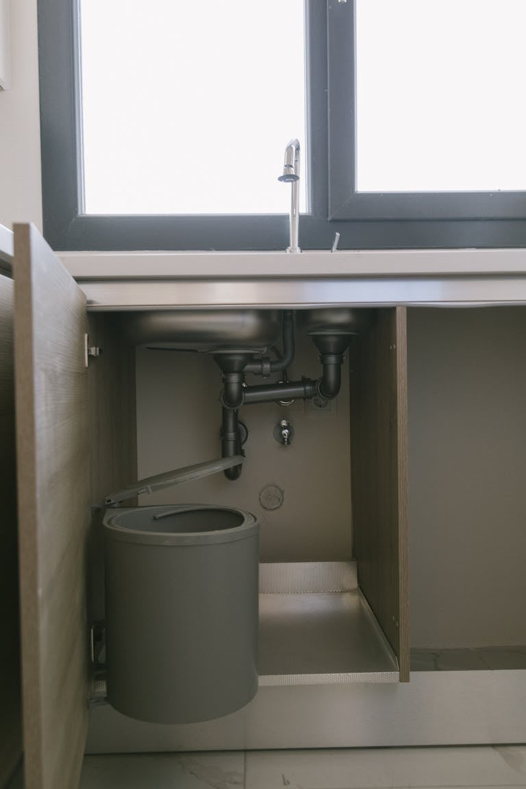 Under-sink view of modern plumbing with garbage bin in an Istanbul apartment kitchen.